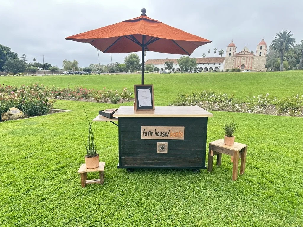 A mobile food stand with a brown umbrella, situated on a grassy lawn with a garden and a historic building in the background. The stand has signage reading 'farm house bento', with small wooden tables and potted plants nearby.