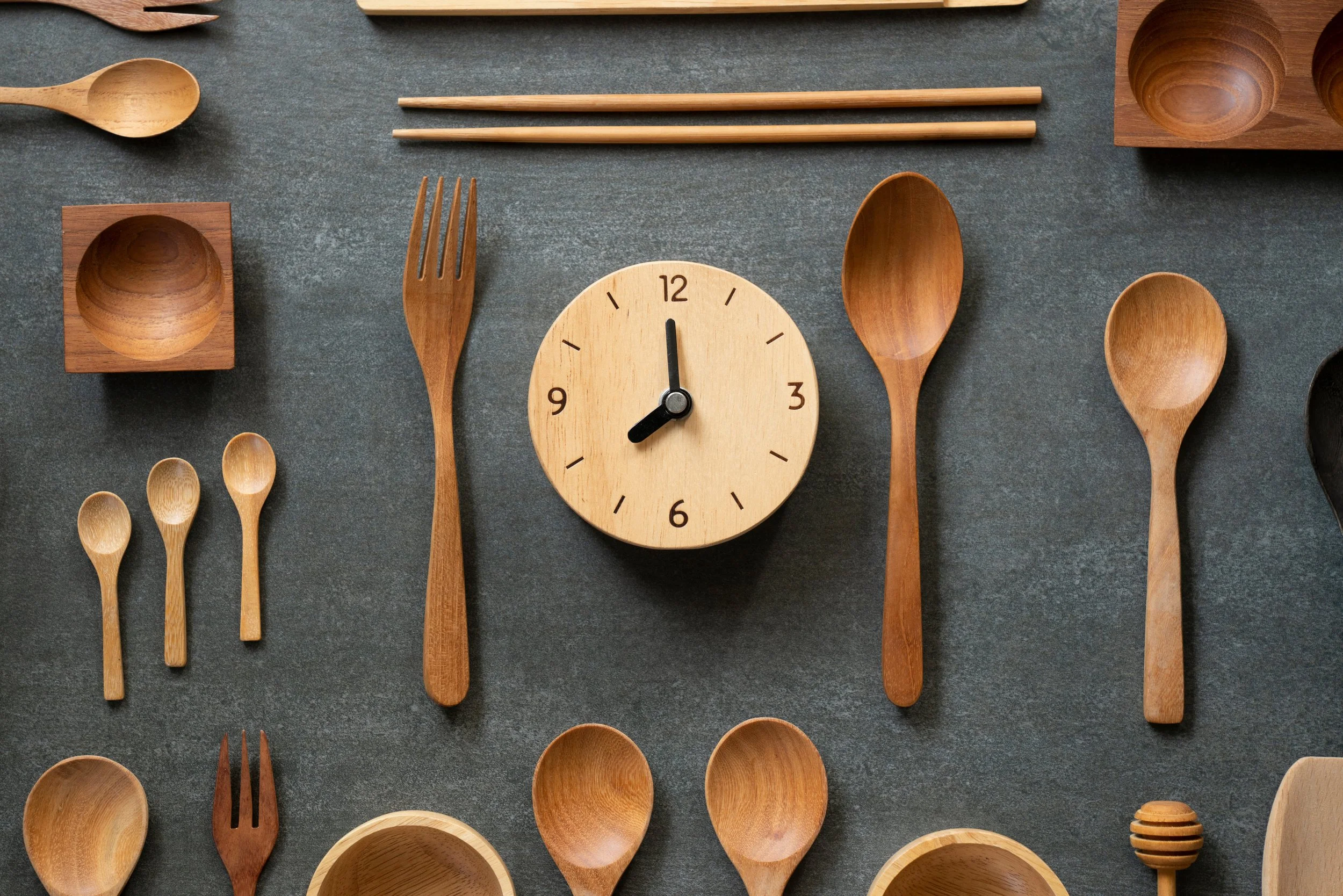 A collection of wooden kitchen utensils and a clock arranged on a dark surface.