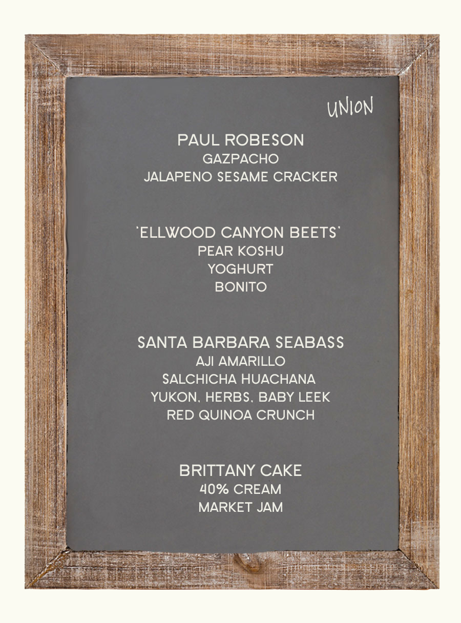 Menu board with a wooden frame listing dishes, including Paul Robeson, Ellwood Canyon Beets, Santa Barbara Seabass, and a Brittany Cake, arranged in sections with various ingredients and descriptions.