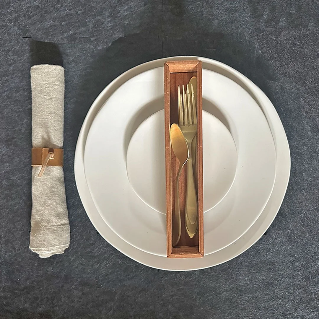 A place setting with a white plate, a wooden holder containing a fork and spoon, a rolled cloth napkin, and a dark textured surface background.