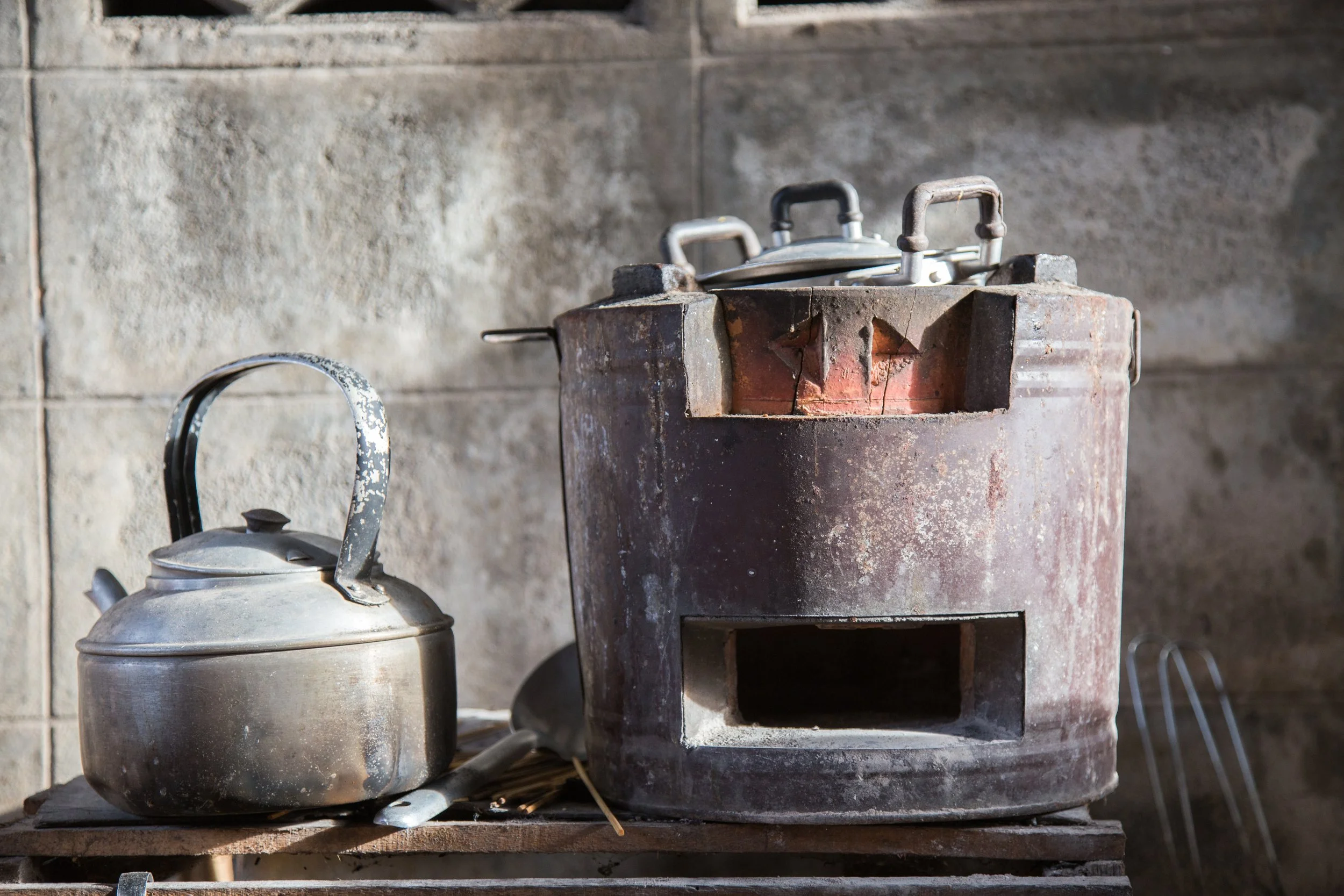 An old, rusty stove with a teapot on the left and an iron on top of the stove against a concrete wall.