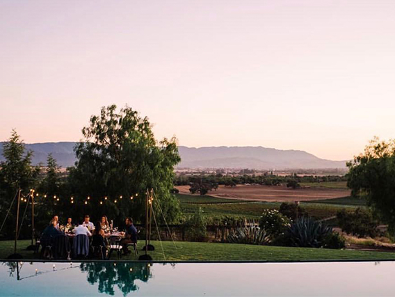 People sitting around a table outdoors near a pool, with string lights overhead and a scenic landscape of fields and mountains in the background during sunset.