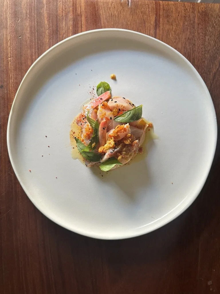 A plate of thinly sliced fish, fresh basil leaves, and garnishes, possibly served as a seafood appetizer.