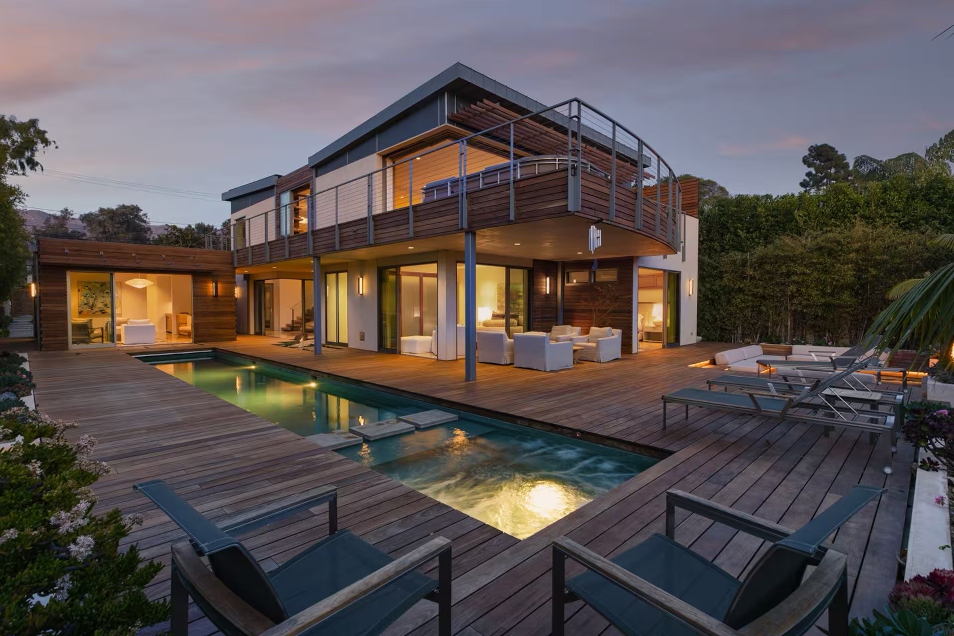 Modern two-story house with large windows, wooden balcony, outdoor seating area, and a pool, surrounded by trees at dusk.