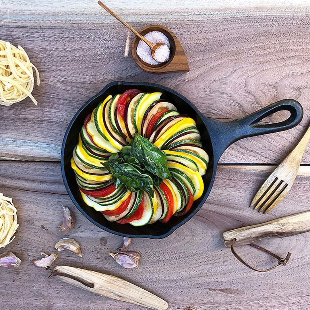 Colorful zucchini and tomato slices arranged in a circular pattern in a black skillet, garnished with basil leaves. Surrounding items include garlic cloves, pasta, a small bowl of salt with a spoon, and wooden utensils on a wooden surface.