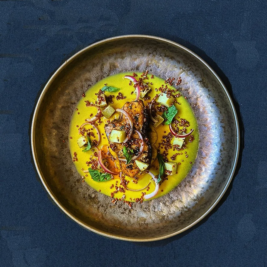 A bowl of yellow curry with grilled fish, garnished with herbs, sliced onions, and red pepper seeds, served in a dark-colored bowl on a dark background.