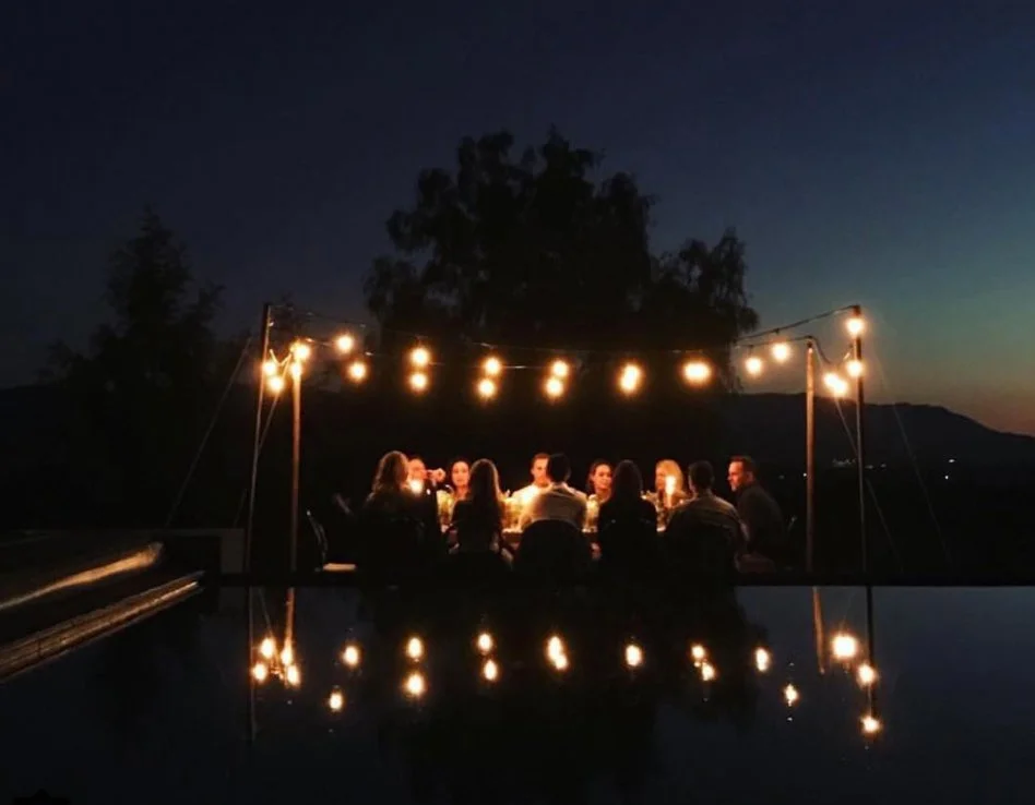 Group of people dining outdoors at night under string lights with a scenic mountain view in the background.