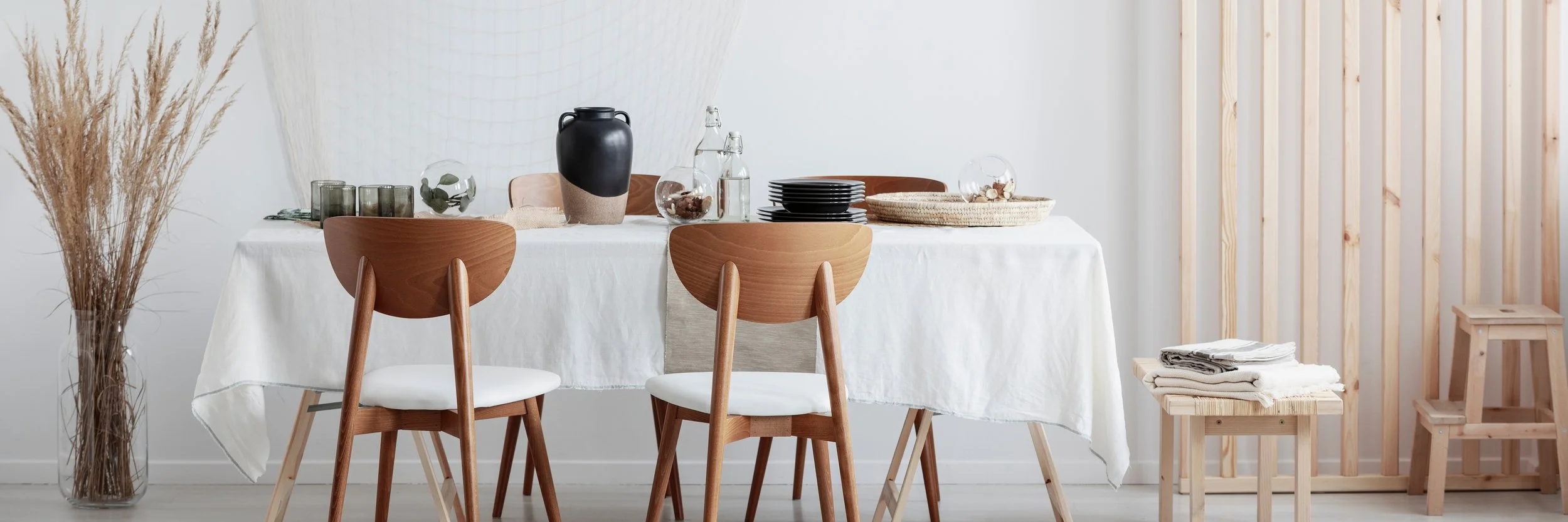 Dining room with a white tablecloth-covered table set with black plates, glassware, and decorative items; wooden chairs and stools, and a tall vase with dried plants against a wall with vertical wooden slats.