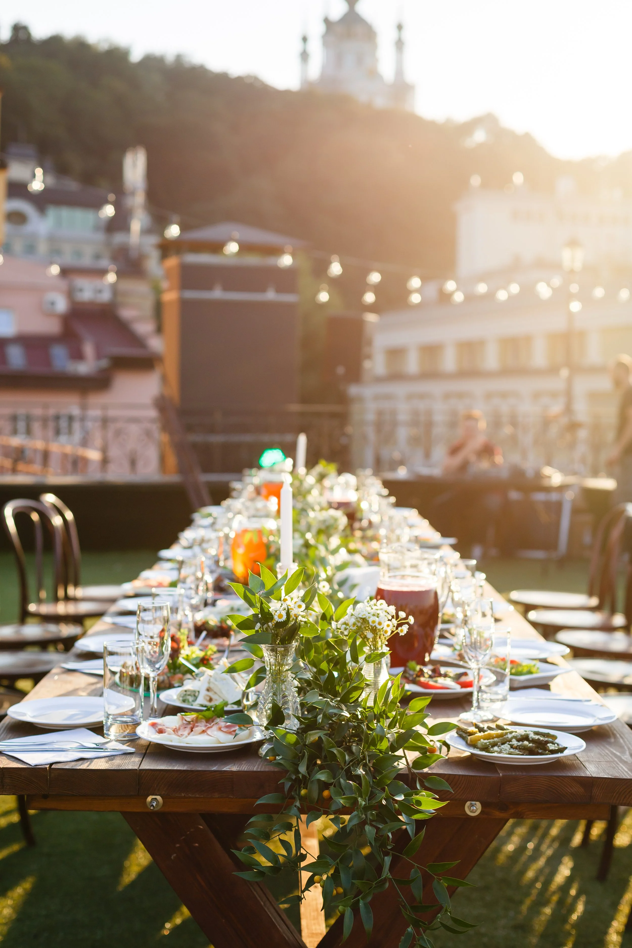 Outdoor banquet table decorated with flowers, candles, and glassware, set for a meal during sunset, with buildings and a hill in the background.