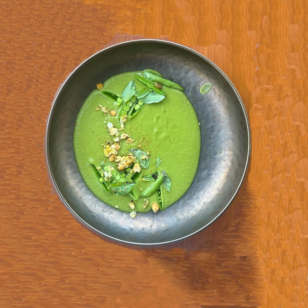 Green soup garnished with herbs, chopped vegetables, and possibly nuts, served in a black bowl on a wooden table.