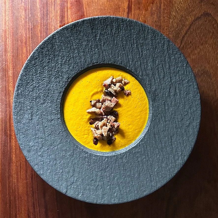A bowl of bright yellow soup garnished with chopped cooked meat and possibly black beans, placed on a brown wooden surface.
