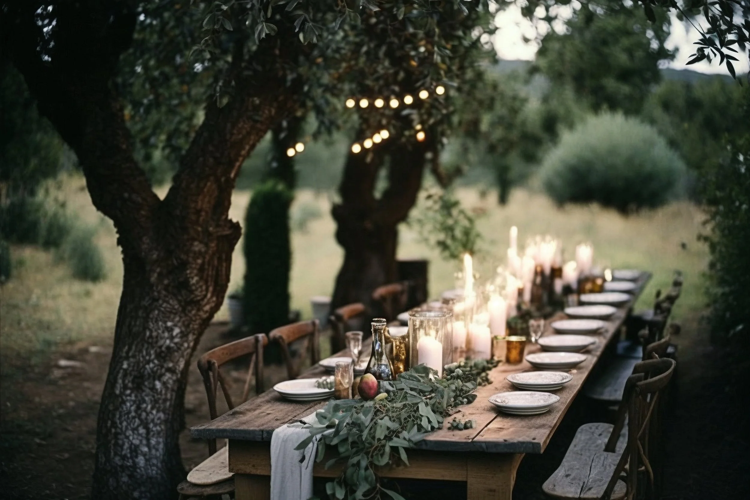 A long outdoor dining table decorated with candles, greenery, and apples, set amidst trees for an evening gathering. String lights hang from the trees above, creating a warm ambiance.