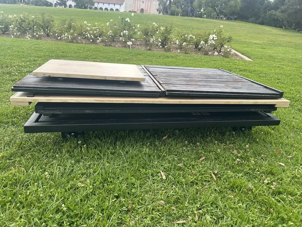 Stack of black and light-colored folding wooden tables on grass in a park with flowers and a building in the background.