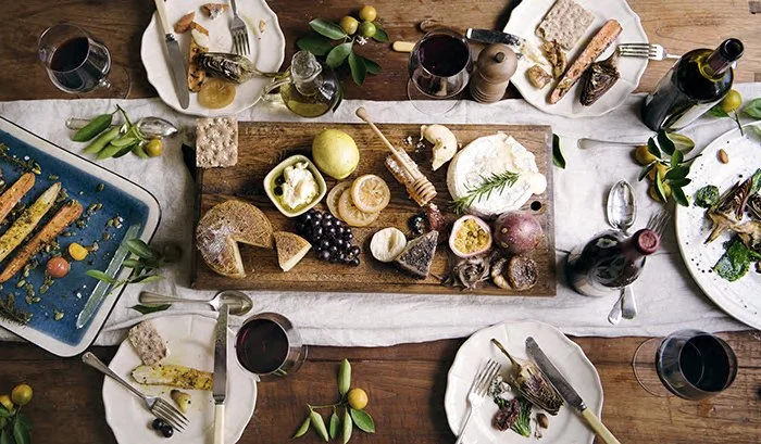 A rustic dinner table set with cheese, fruit, bread, and wine, with plates, glasses, and utensils arranged around it.
