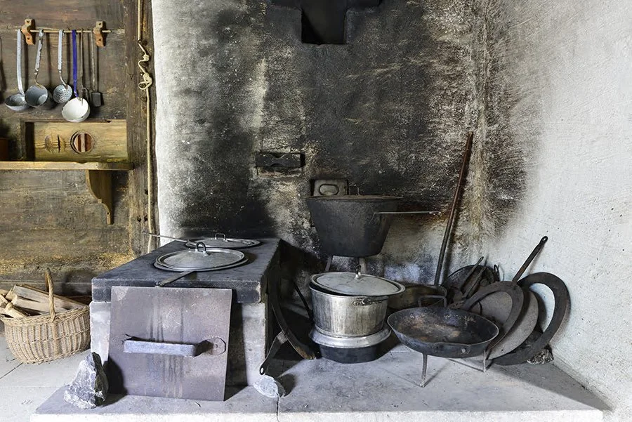 Old kitchen scene with a wood-fired stove, cast iron cookware, and hanging utensils, in a rustic setting.