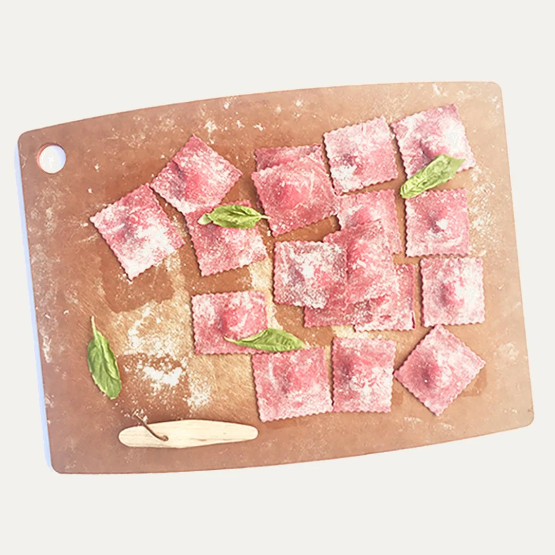 Fresh square-shaped ravioli pasta with a pink filling on a floured wooden cutting board, garnished with basil leaves.