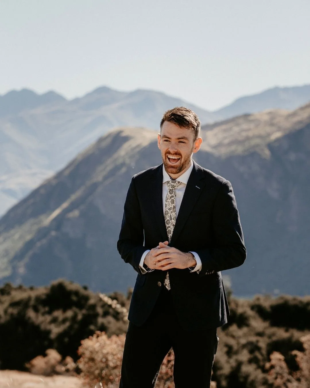 A man smiling outdoors in a snowy mountainous area, wearing a beige suit, a white shirt, and a colorful bow tie, holding a laptop.