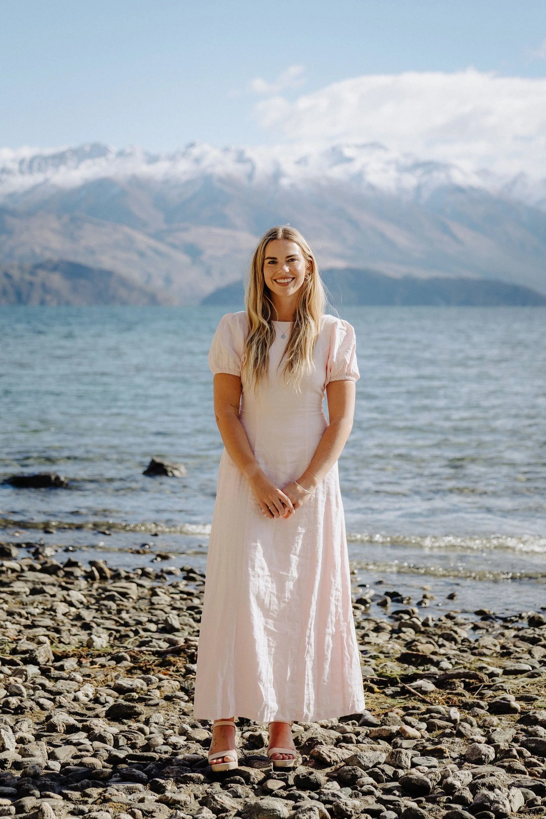 A woman in a long, pale pink dress standing on a rocky shoreline with mountains and a lake in the background.