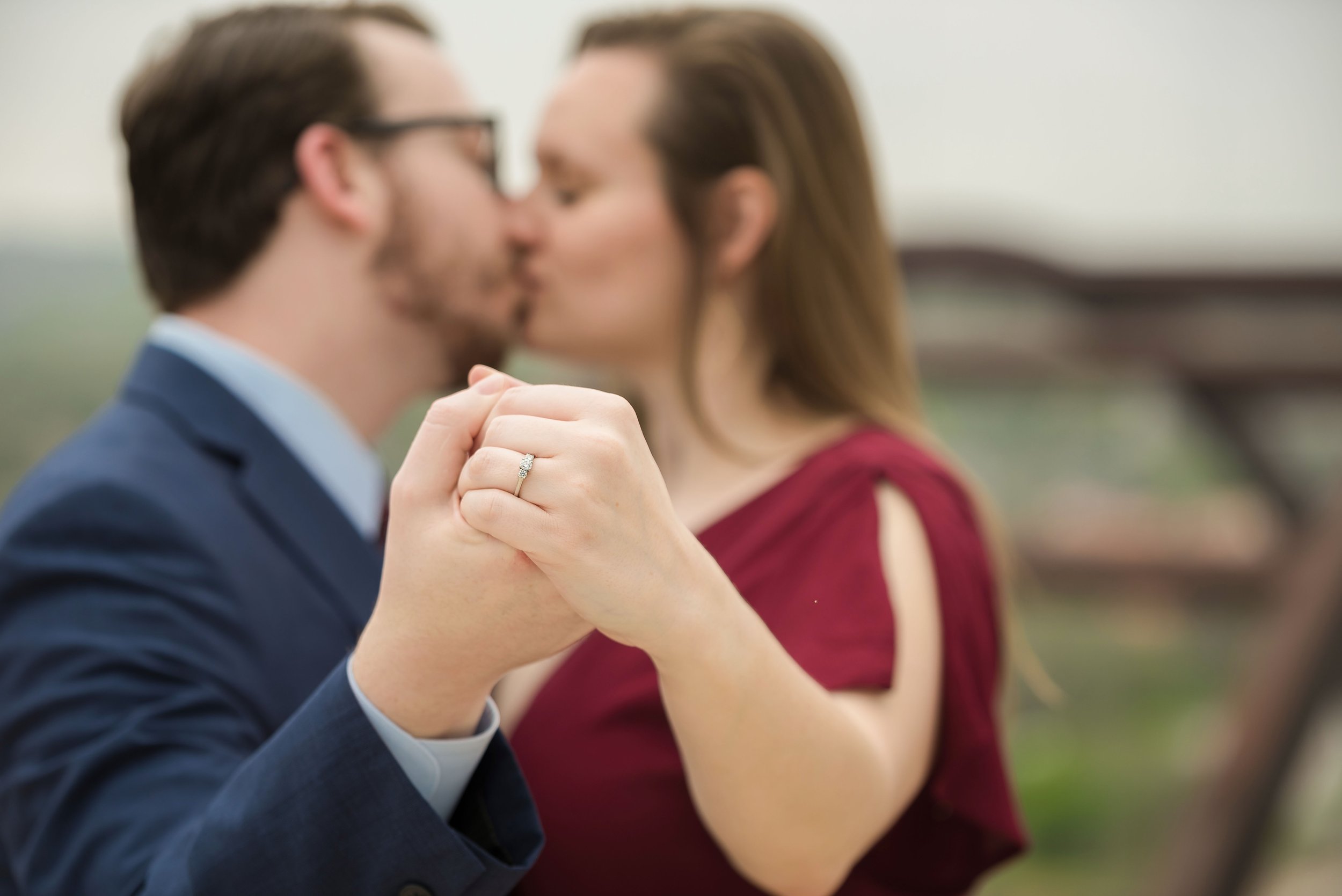 A couple kissing, holding hands with a wedding ring visible, outdoors in a park.