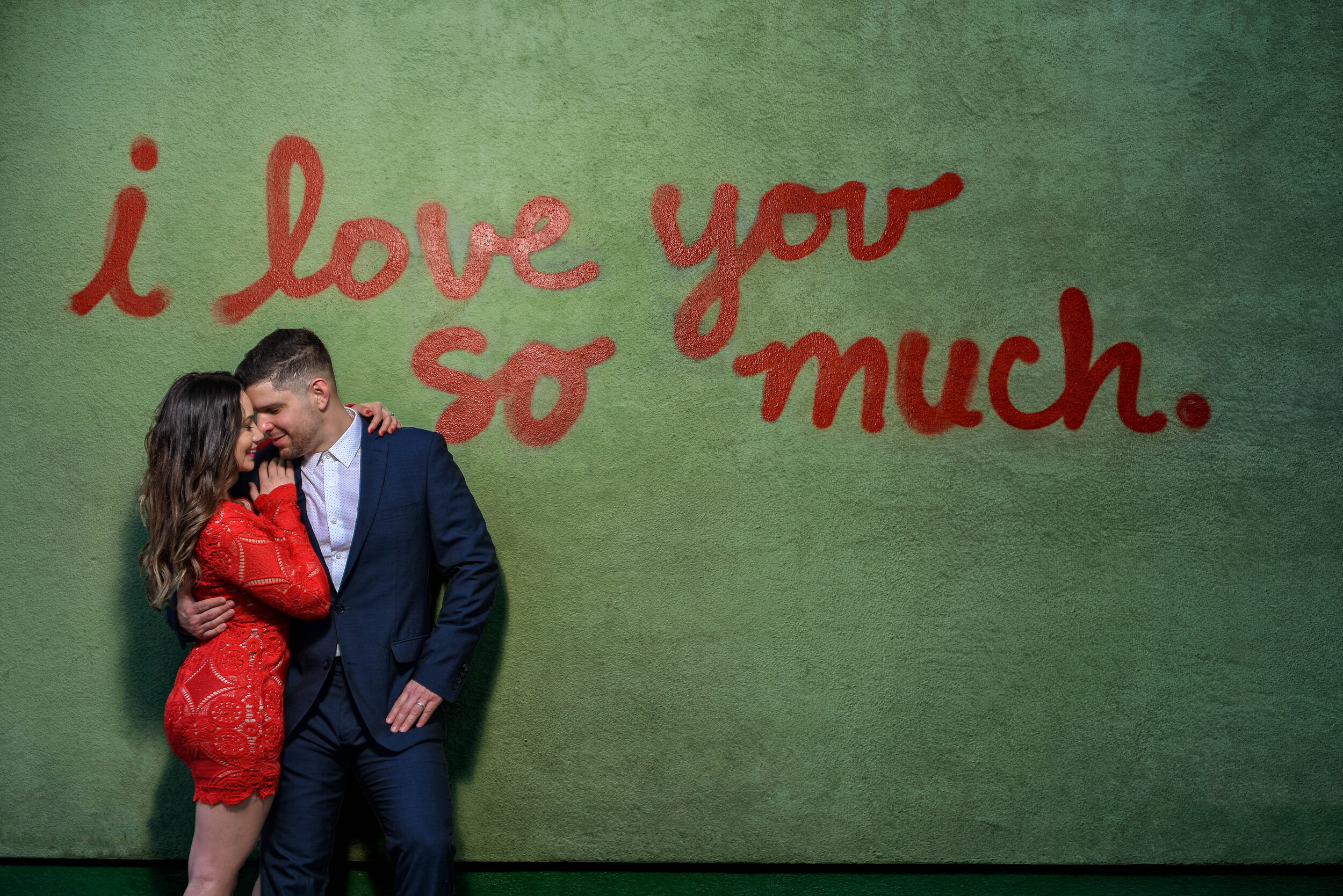 A couple embracing against a green wall with the phrase 'i love you so much' written in red letters.