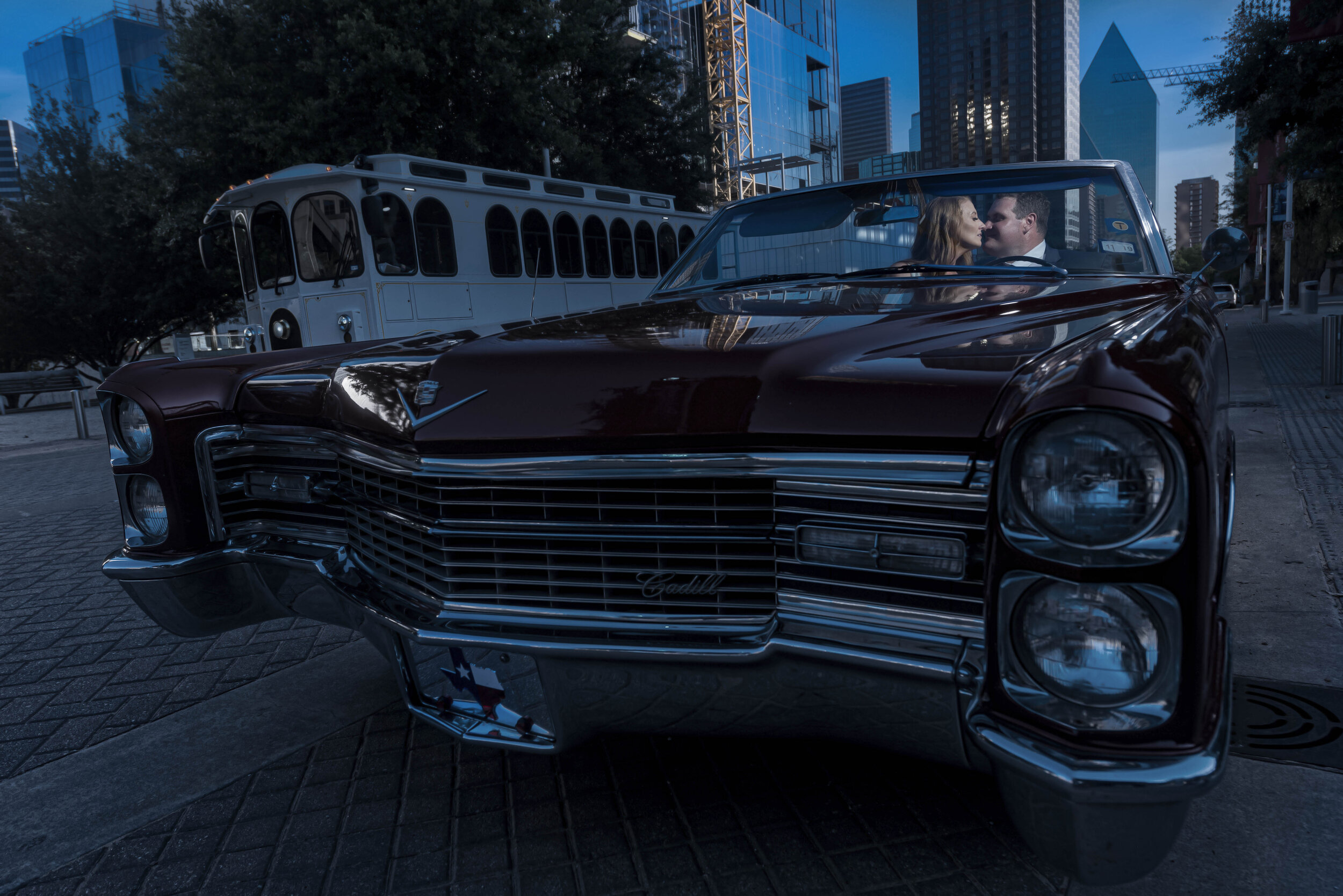 A vintage black Cadillac convertible with a couple inside, parked on a city street at dusk with modern tall buildings and a trolley in the background.