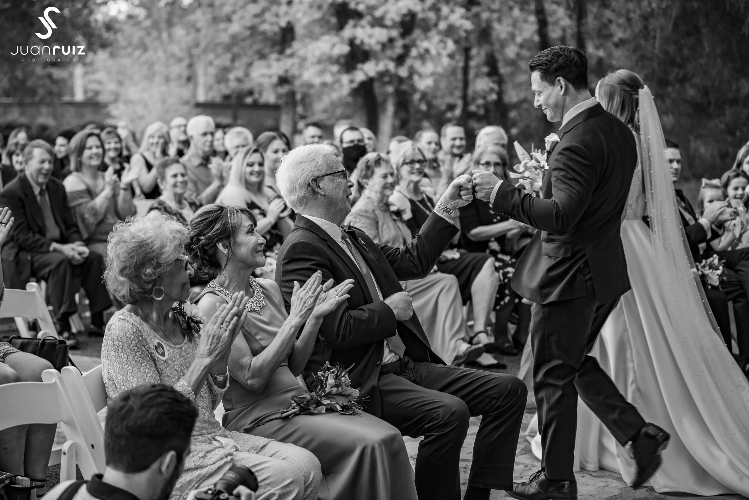 Black and white photo of a wedding ceremony outdoors, showing the bride and groom exchanging rings in front of seated guests. The groom is in a suit, and the bride’s veil is flowing behind her. Guests are clapping and smiling.