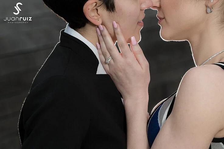 Close-up of a couple about to kiss, with the woman gently holding the man's face. The woman is wearing a dress with a black, white, and blue pattern and a pearl necklace, while the man is dressed in a tuxedo or suit. The background is blurred.