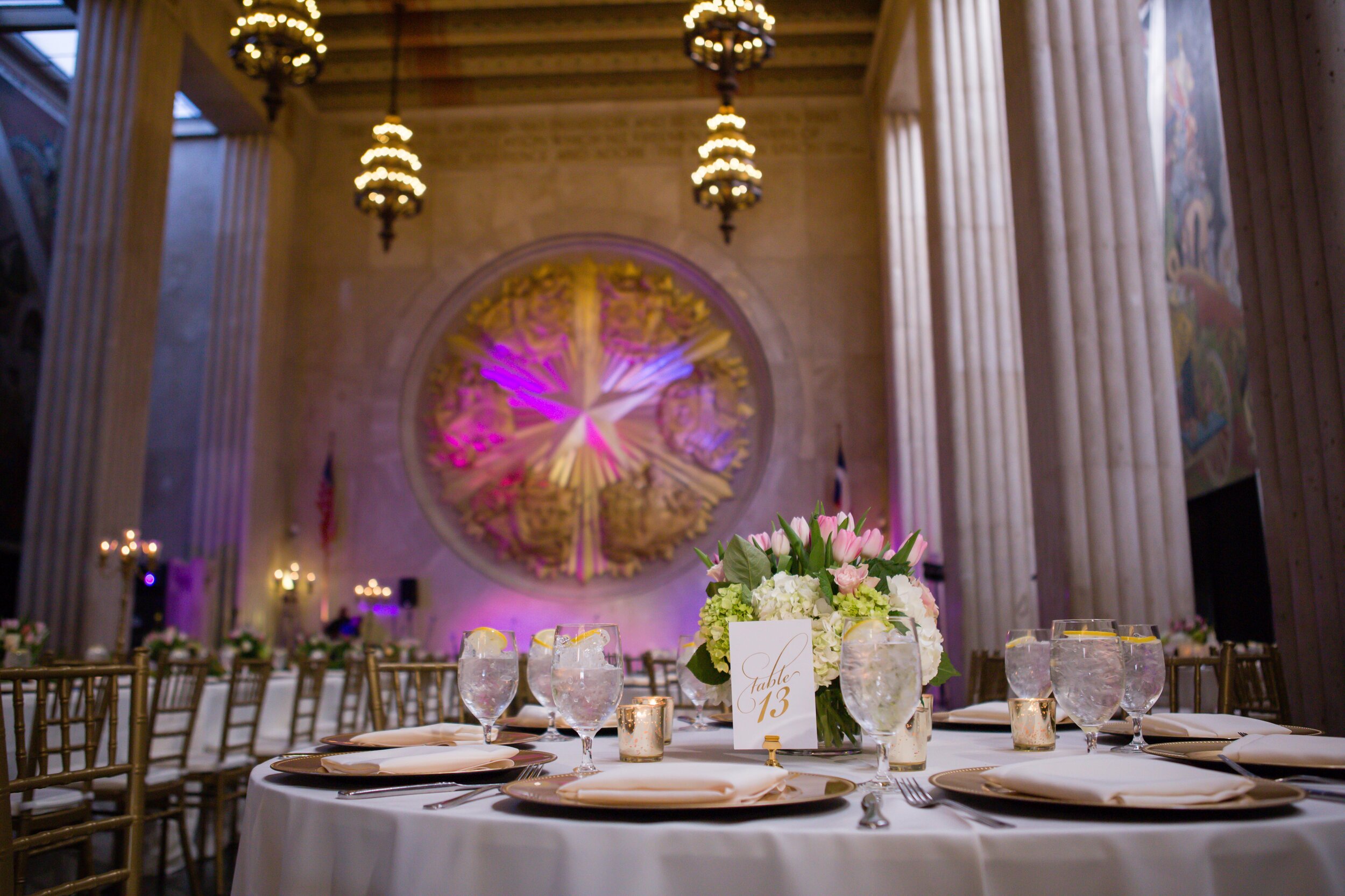 Elegant banquet table set for a formal event with floral centerpiece labeled 'Table 13', surrounded by gold chairs, with a large decorative clock on the wall and tall columns in the background.