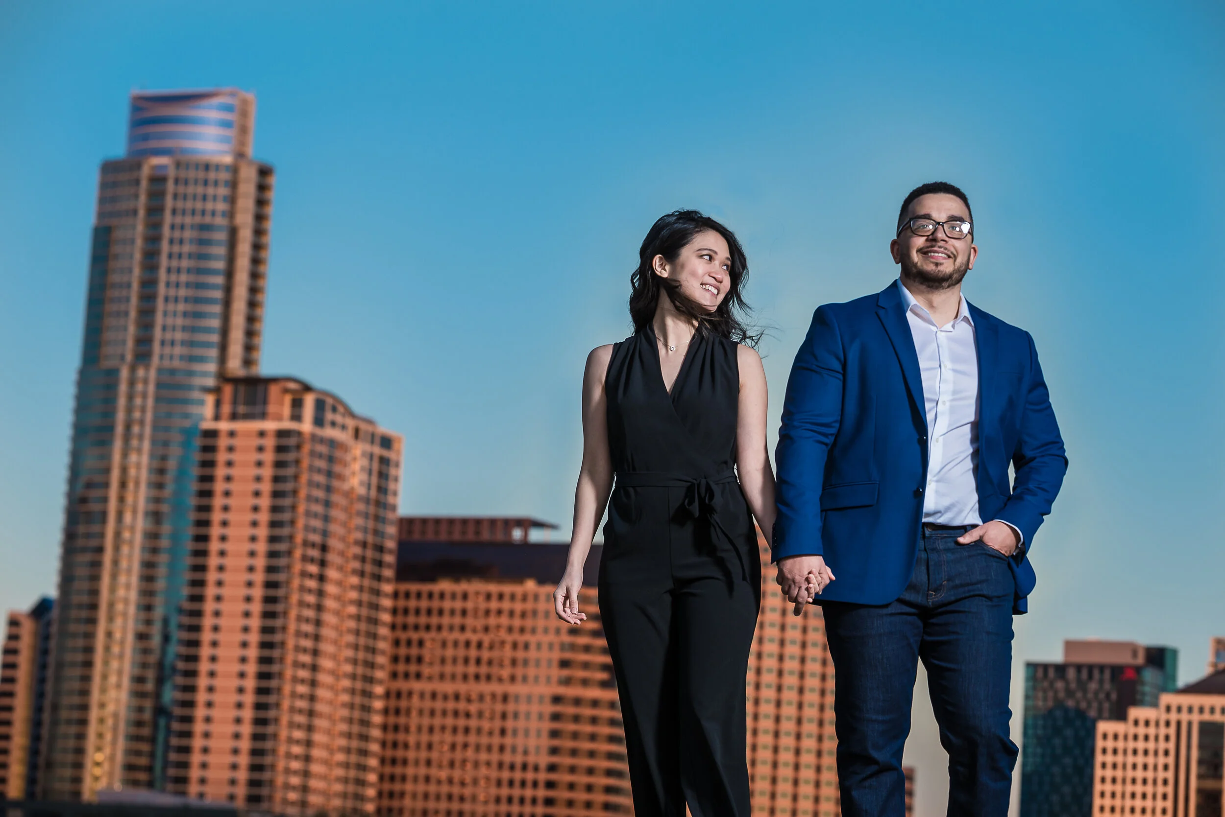 A man and woman holding hands and walking outdoors in front of city buildings, with the woman looking at the man and smiling, both dressed in business attire.