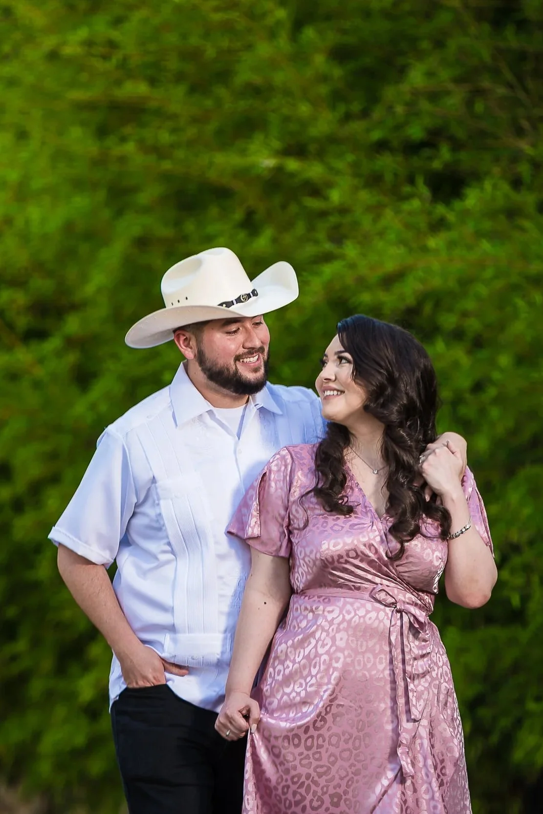 A smiling couple stands close together outdoors with green foliage in the background. The man wears a white cowboy hat and a light blue button-up shirt, while the woman wears a pink leopard print dress.