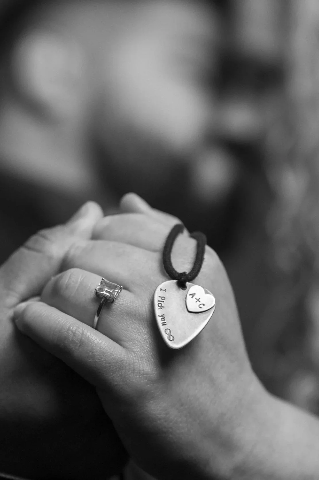 Close-up of a hand with a ring and two engraved heart-shaped and oval charms hanging from a string, with the background blurred.