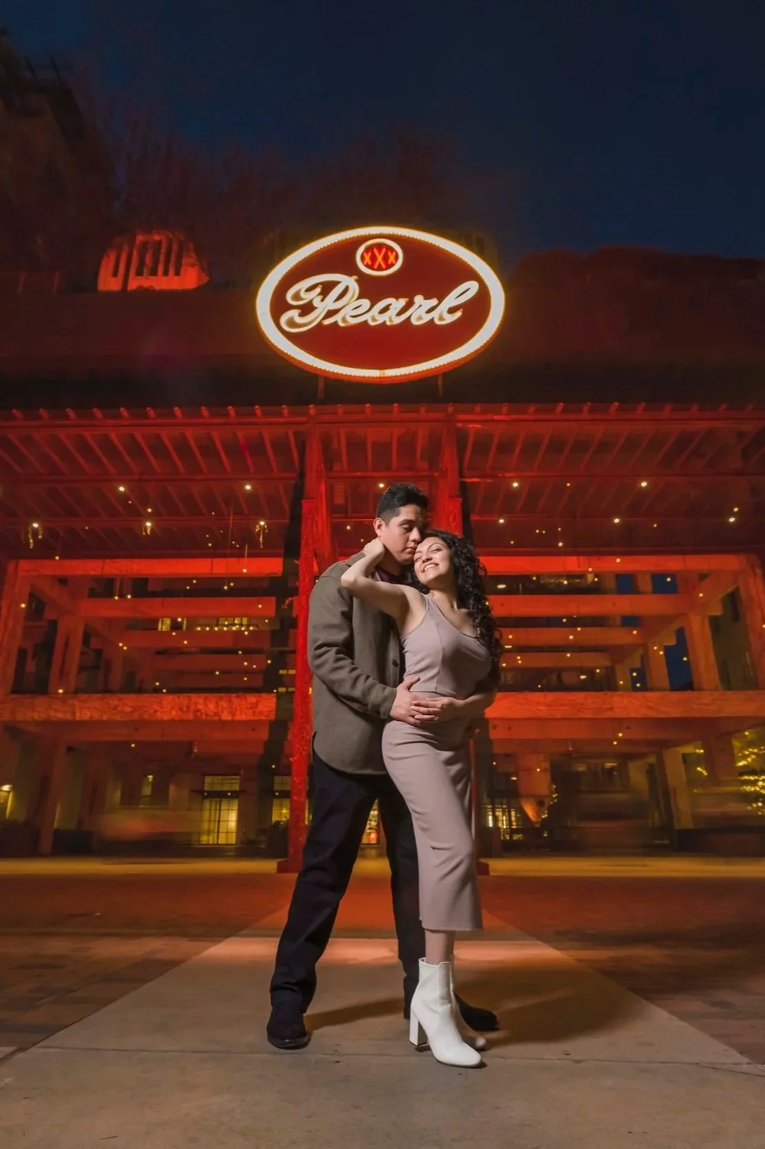 A young couple standing close together outside a building at night, with a red neon sign above that reads 'Pearl' and 'XX', illuminated against a dark sky.
