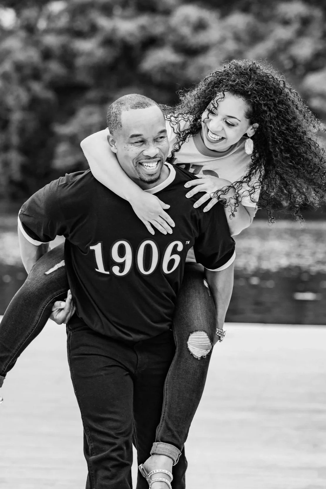 A man carrying a woman on his back outdoors, both smiling and laughing, with a blurred natural background.