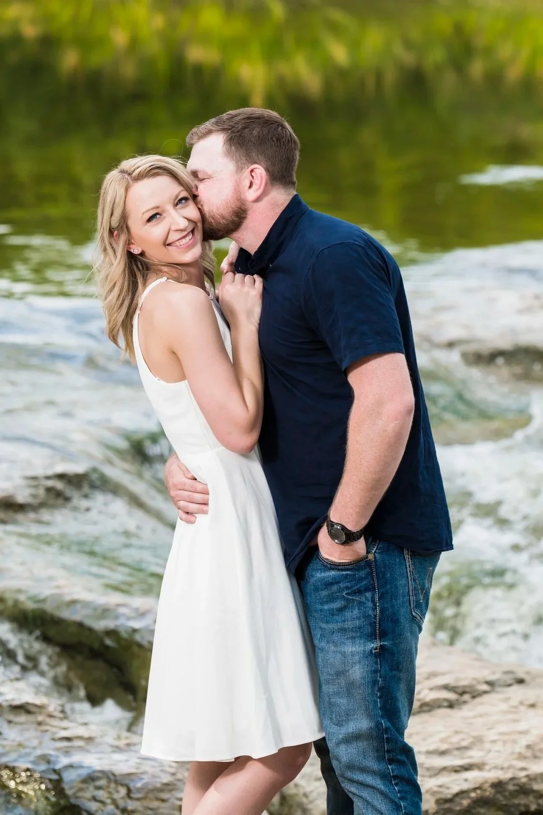 A smiling woman in a white dress is being kissed on the cheek by a man in a dark blue shirt by a river, with greenery in the background.