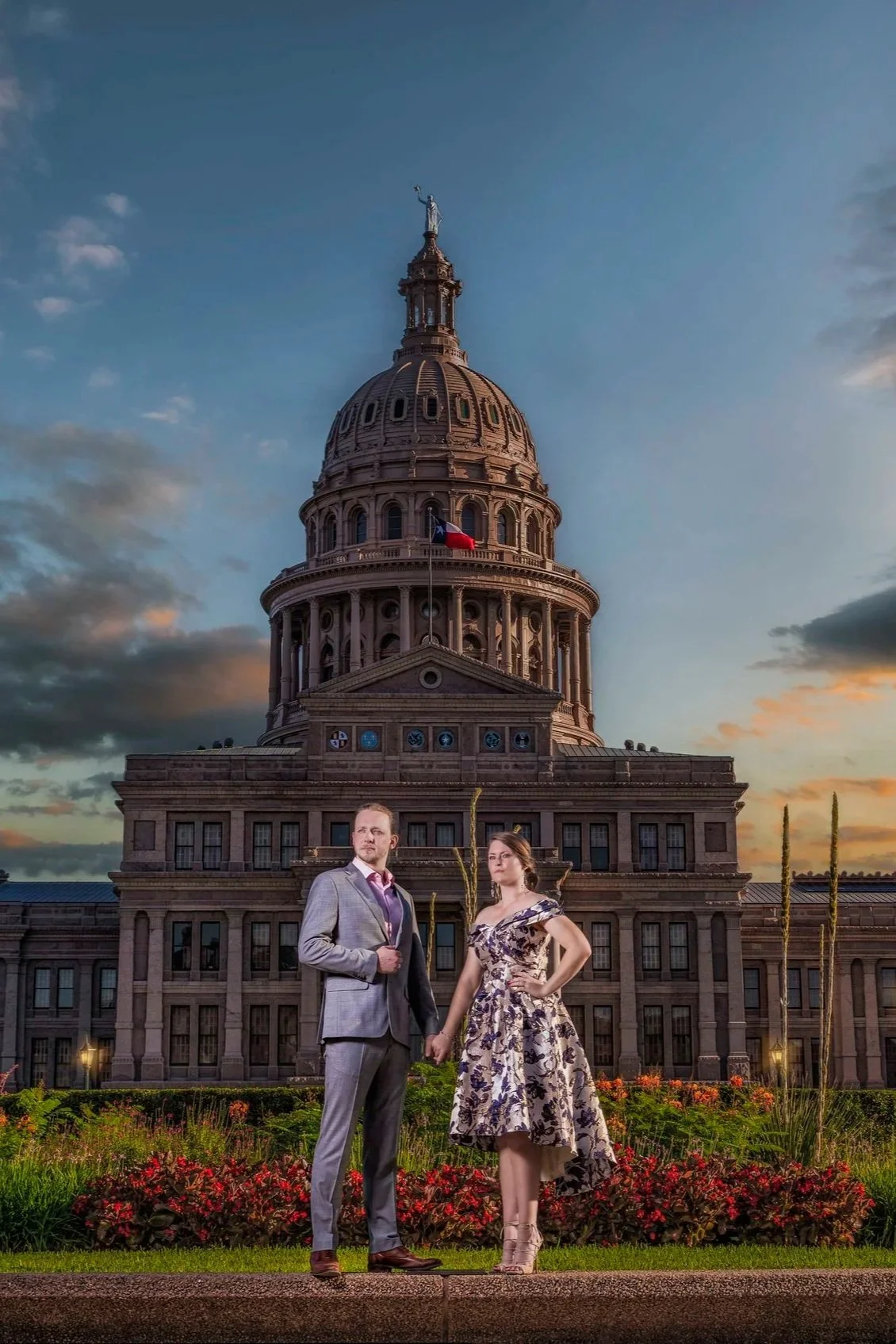 A man and woman standing in front of the Texas State Capitol building at sunset, holding hands, with the woman wearing a floral dress and the man in a suit.