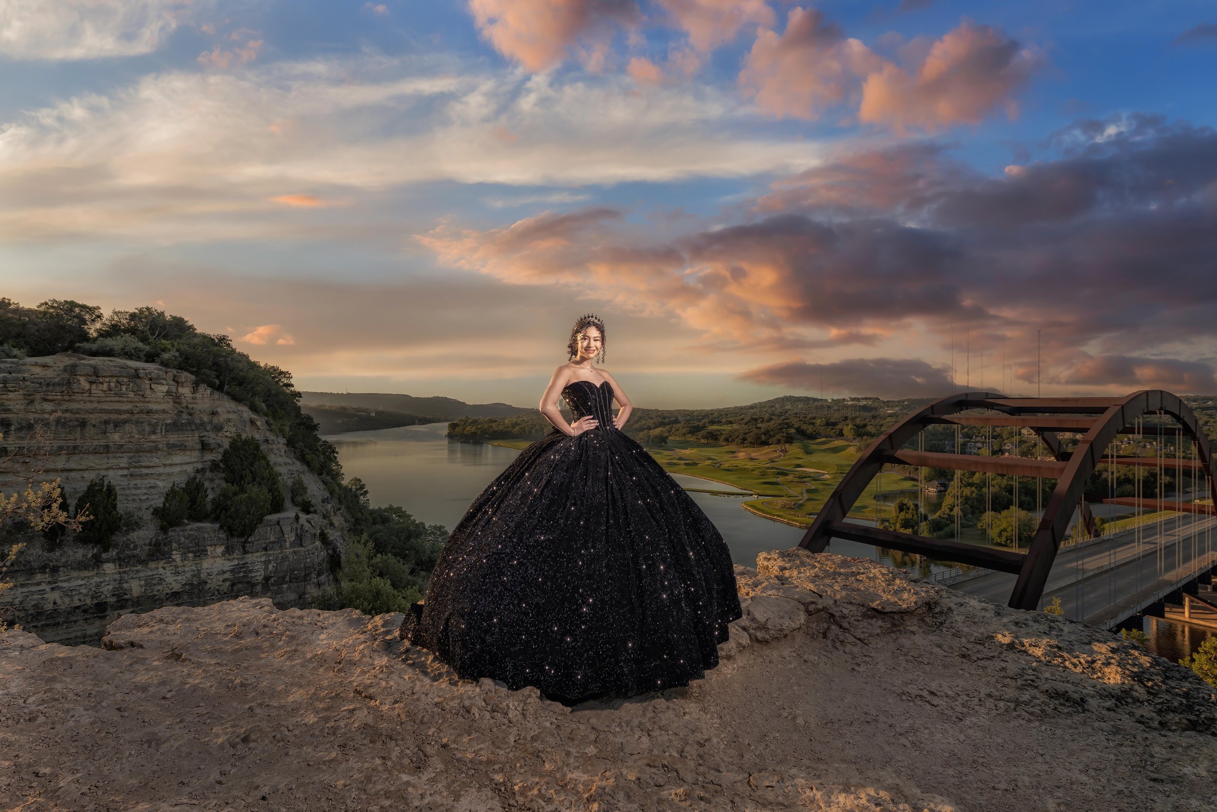 A woman in a black starry ball gown standing on a rocky hilltop at sunset with a river, hills, and a bridge in the background.