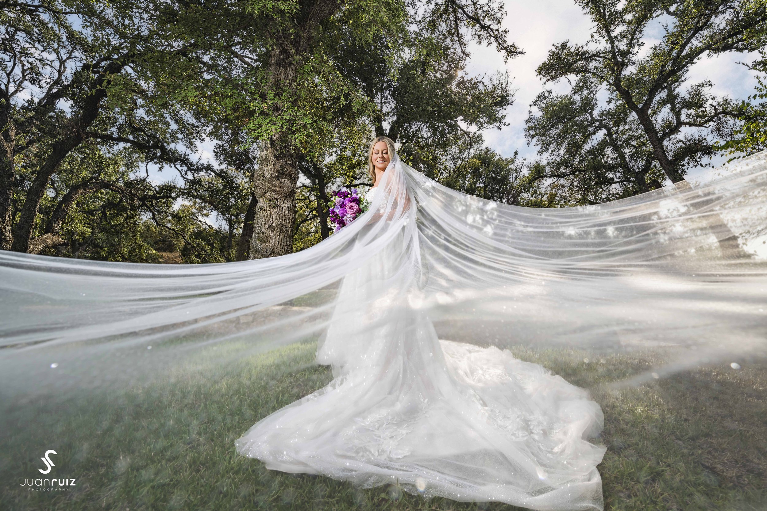 A smiling bride in a white wedding dress and veil holding a bouquet of purple and pink flowers outdoors among trees.
