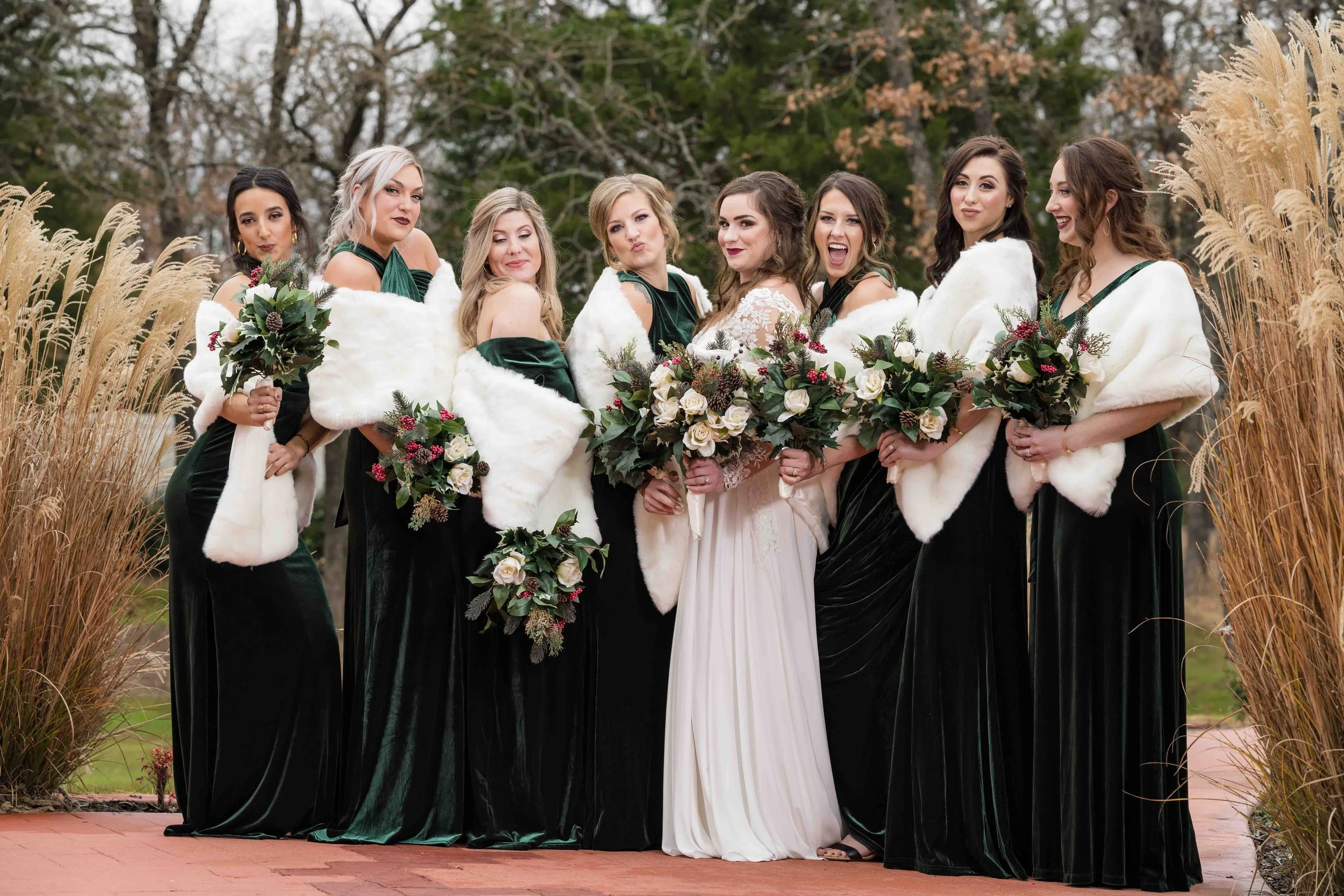 A bride in a white dress holding a bouquet of white and pink roses, surrounded by seven bridesmaids in dark green dresses and white fur shawls, outdoors with tall grasses and trees in the background.