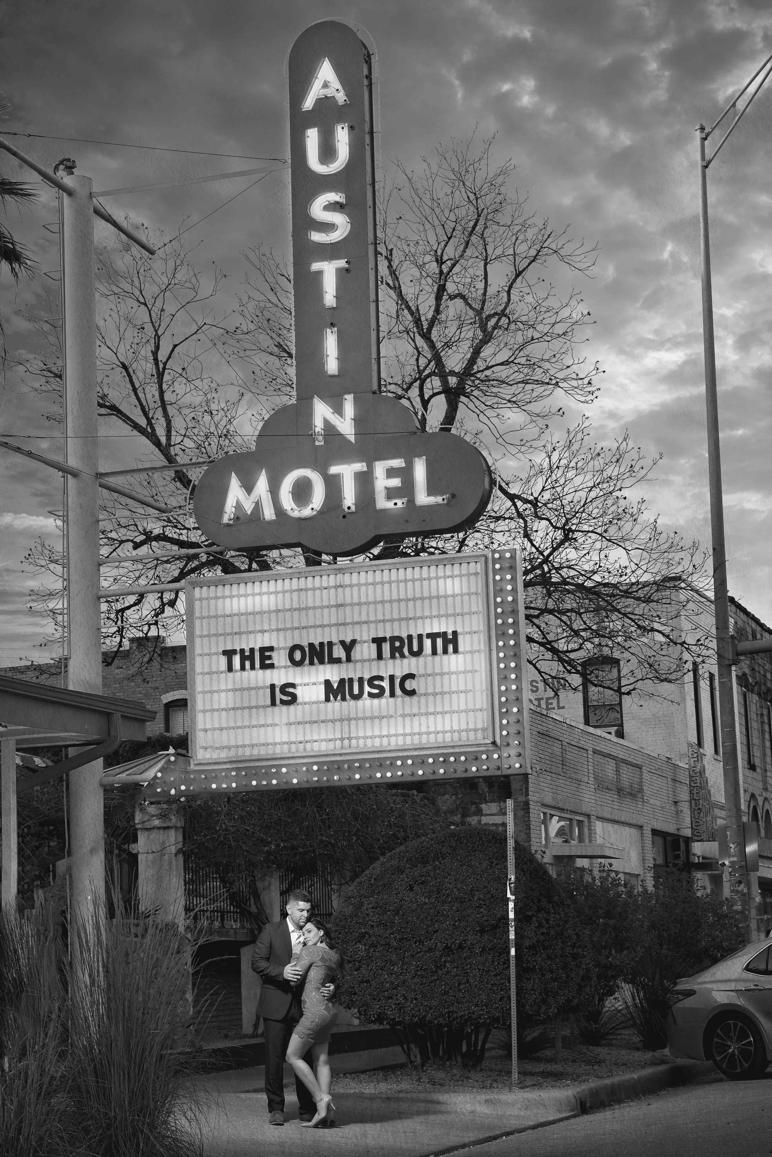 Black and white photo of a vintage motel sign with the words "AUSTIN MOTEL" and a message board that reads "THE ONLY TRUTH IS MUSIC". A man in a suit and a woman in a dress are standing together in front of the sign.