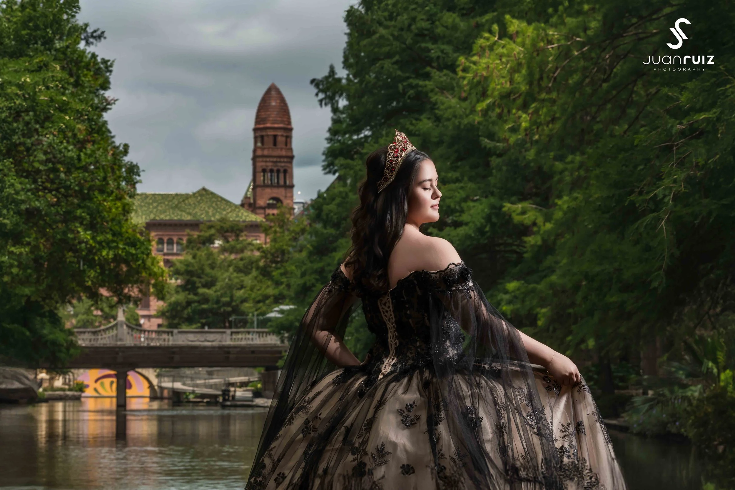 A woman in an elegant black and beige off-shoulder gown with lace details, wearing a tiara, stands by a river with lush green trees, a bridge, and a historic building with a tower in the background.