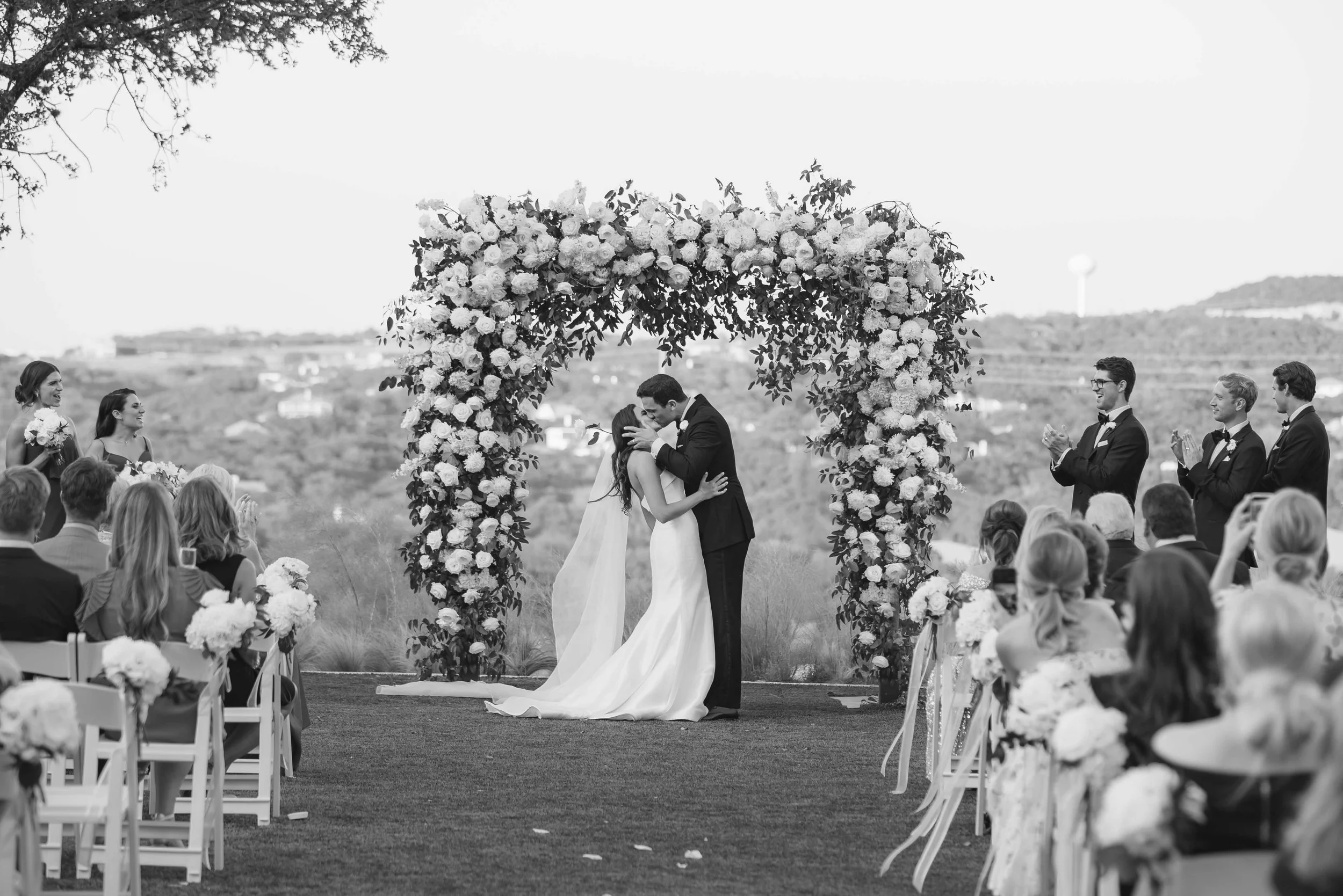 A wedding ceremony outdoors with a bride and groom kissing under a large flower arch, surrounded by guests sitting and standing, with a landscape and water tower in the background.