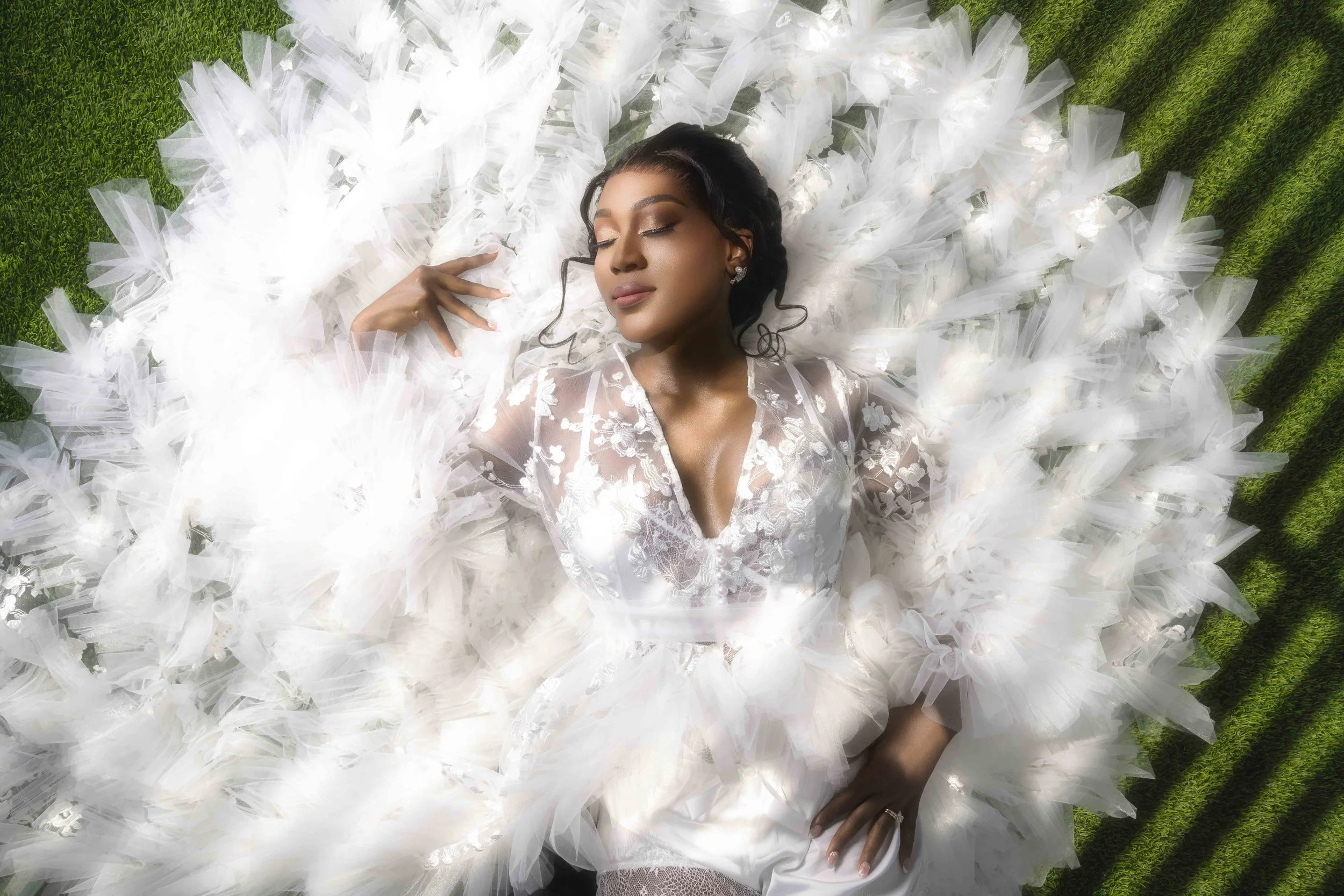 A woman dressed in a white lace gown is lying on green grass, surrounded by white tulle and tulle bows, with her eyes closed and a serene expression.