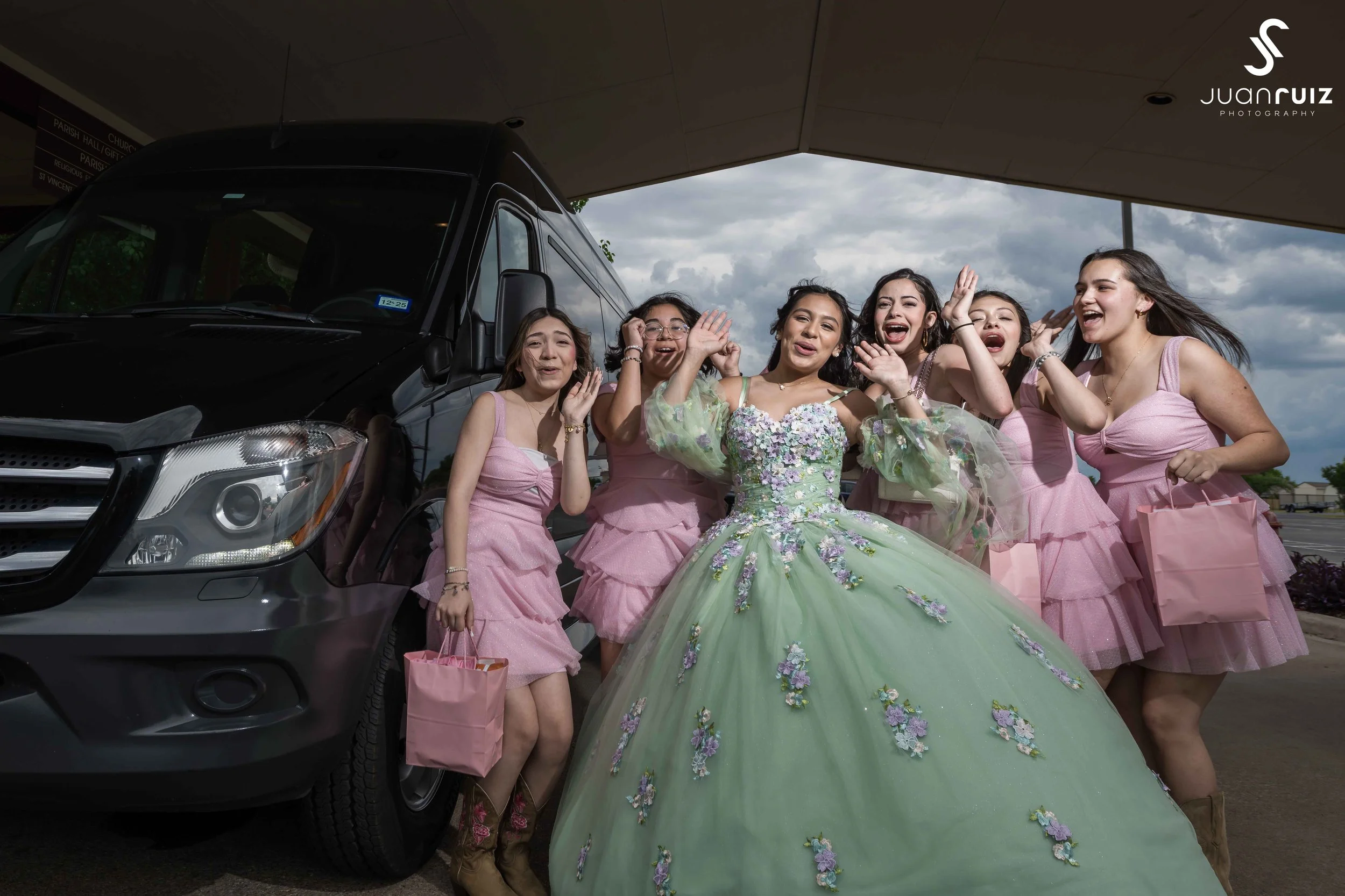 Group of six women, one in a green floral gown and five in matching pink dresses, posing and smiling in front of a black van under a canopy.