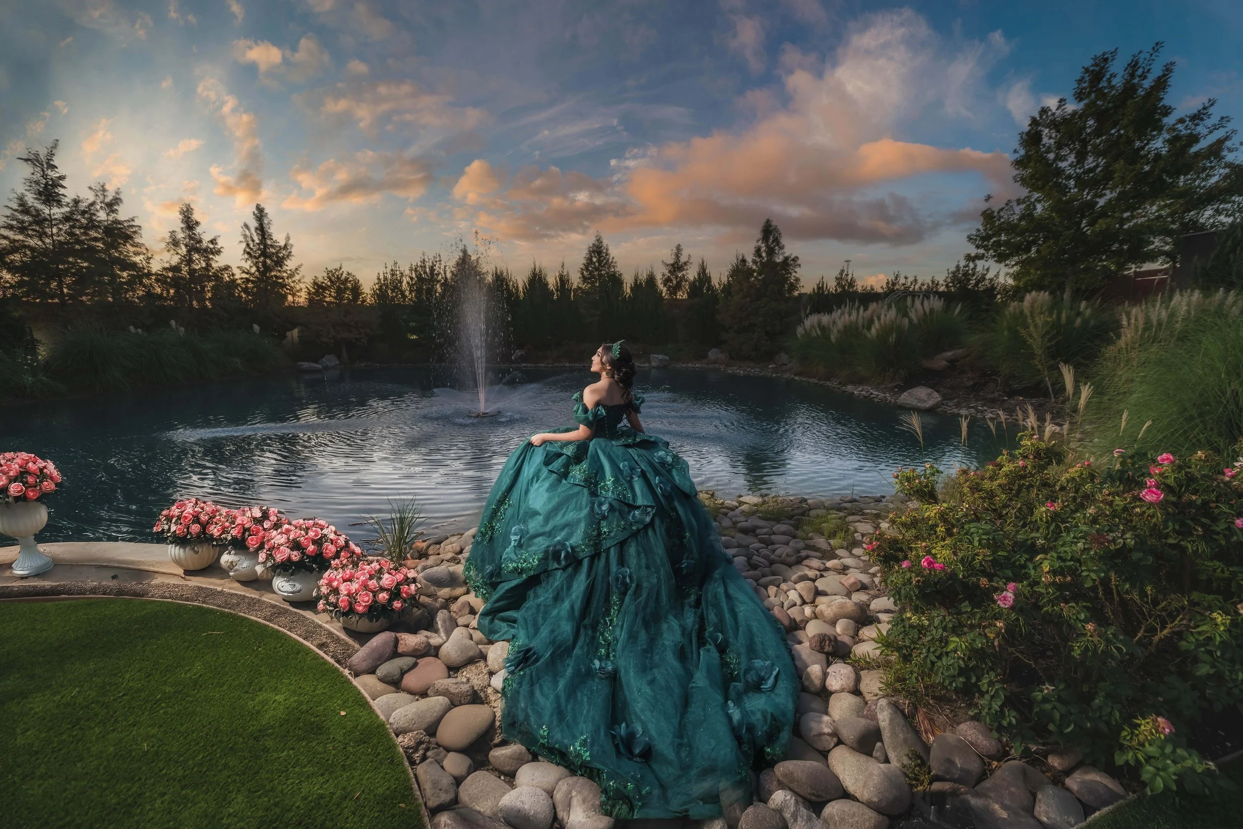A woman in a teal ball gown standing by a pond with a fountain at sunset, surrounded by flowers and trees.