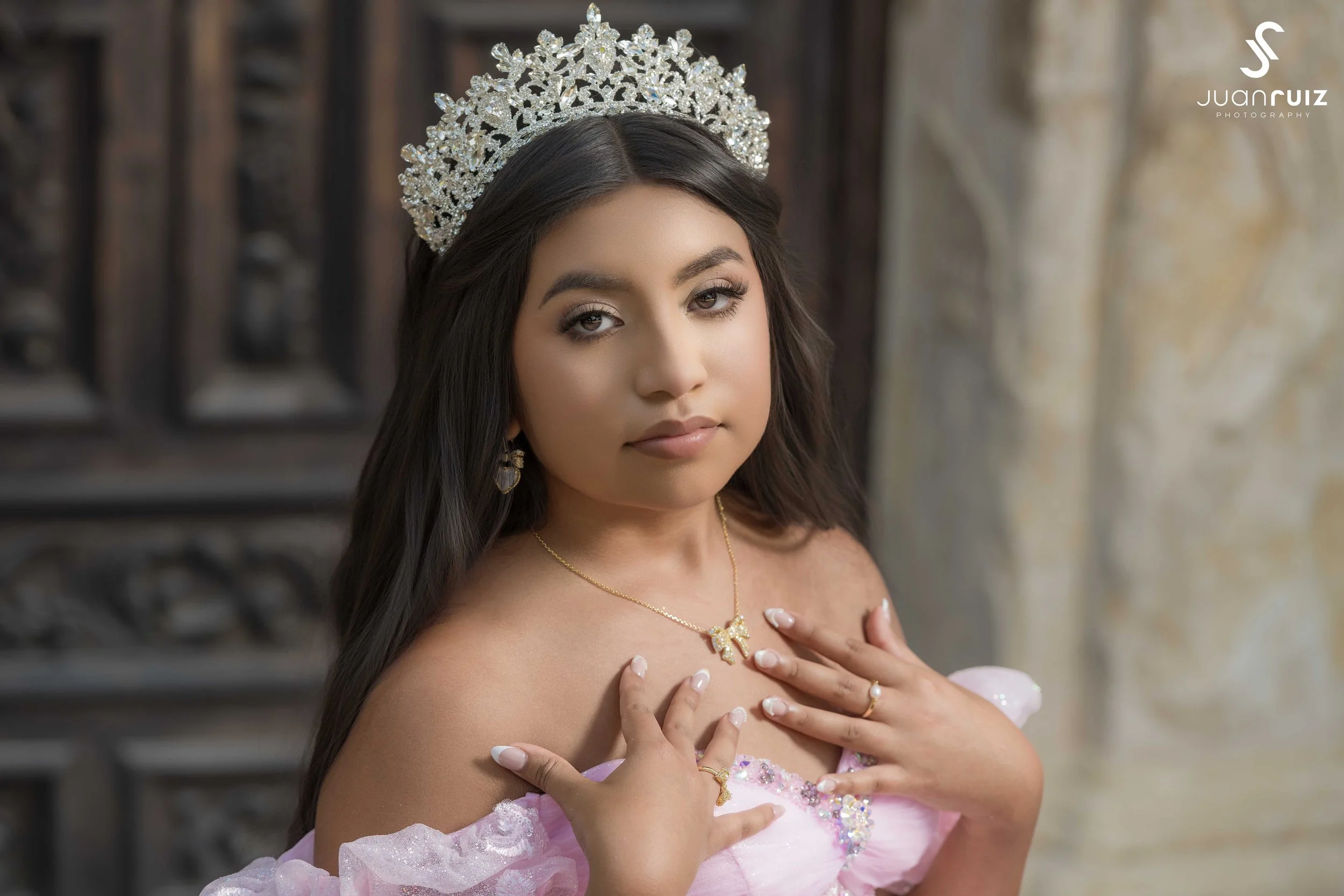 Young woman wearing a silver tiara, pink dress with floral details, and jewelry, posing with one hand on her chest and looking at the camera.