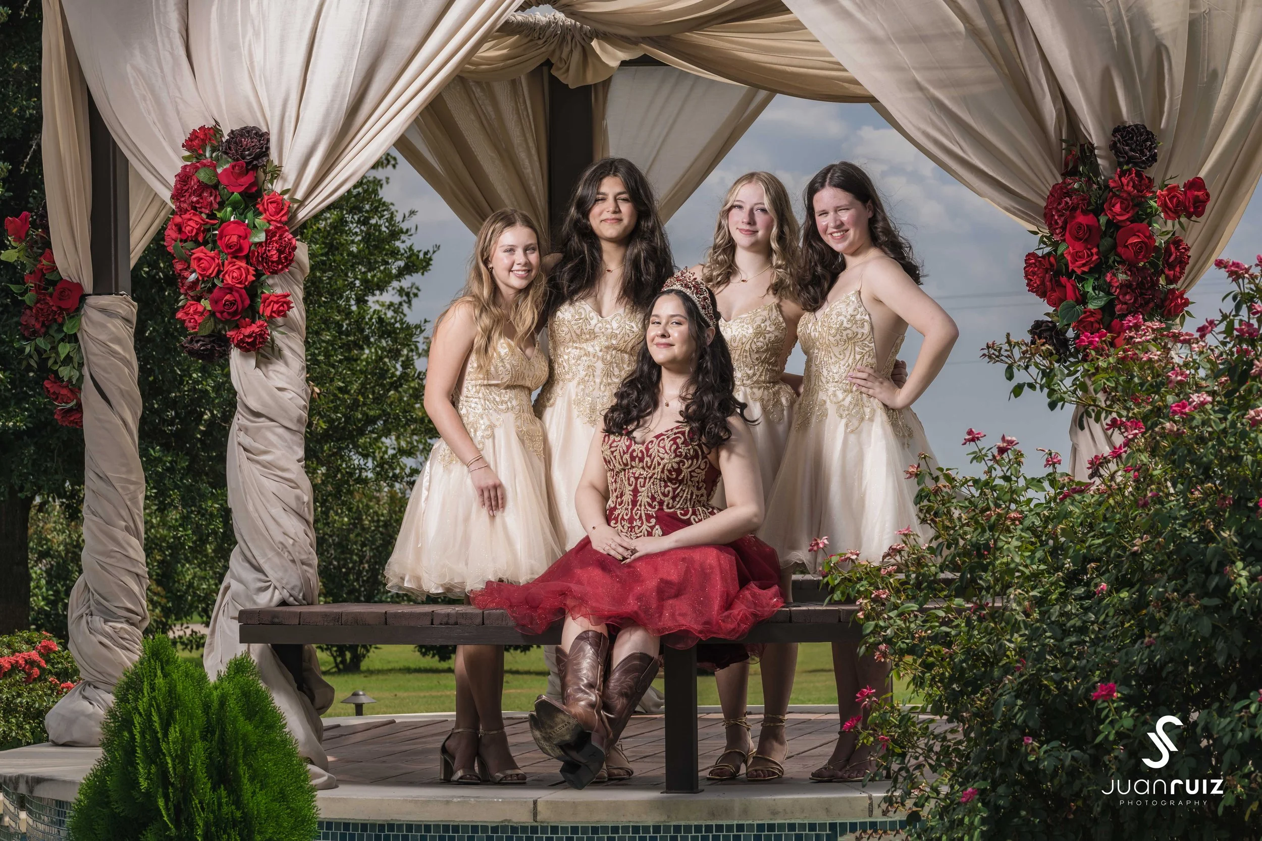 Group of six young women in formal dresses posing under a decorated canopy outdoors, with pink and red flowers, greenery, and a cloudy sky in the background.