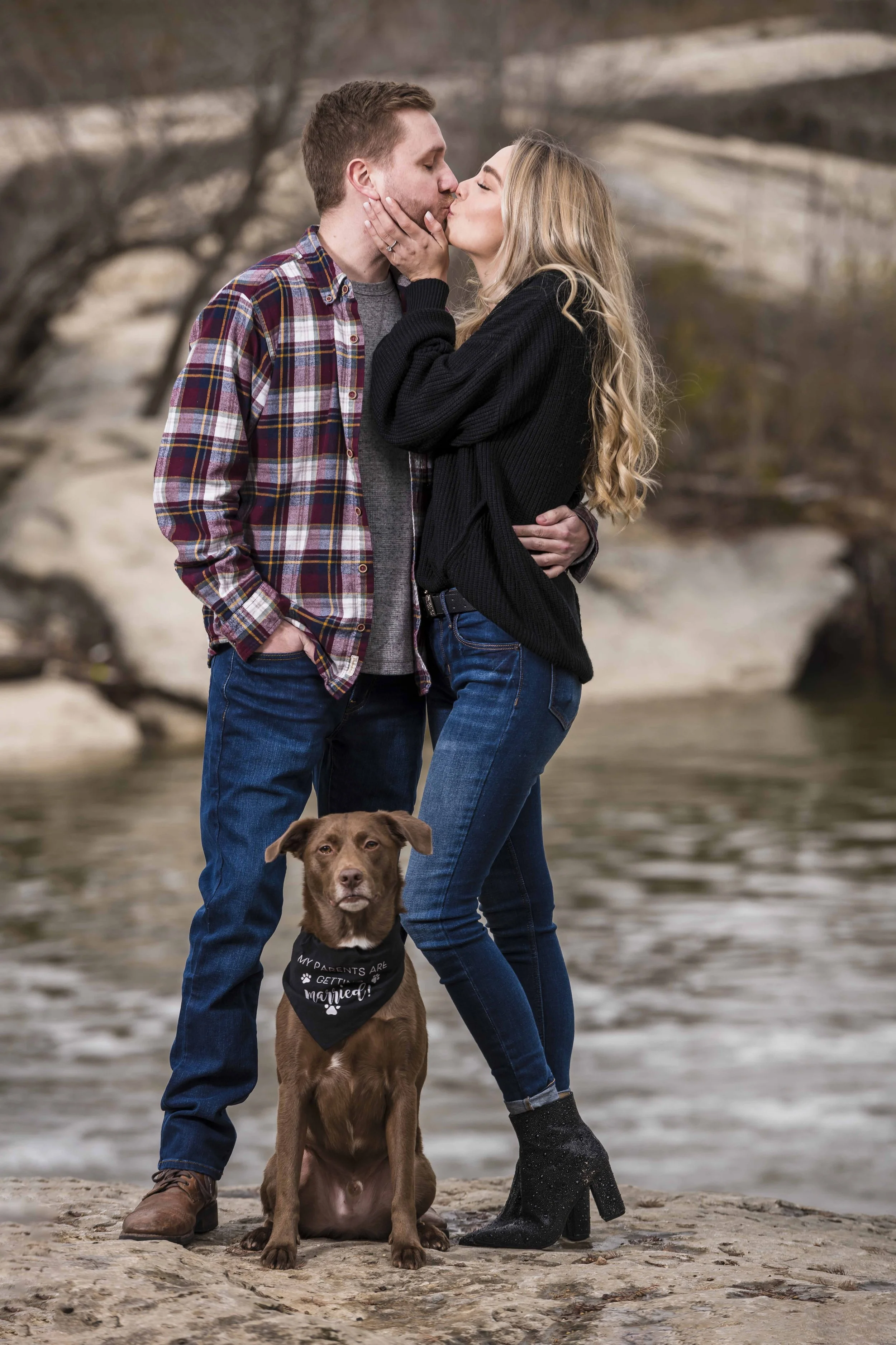 A couple kissing outdoors near a river with a brown dog sitting in front of them, wearing a bandana that says "My parents are getting married!"