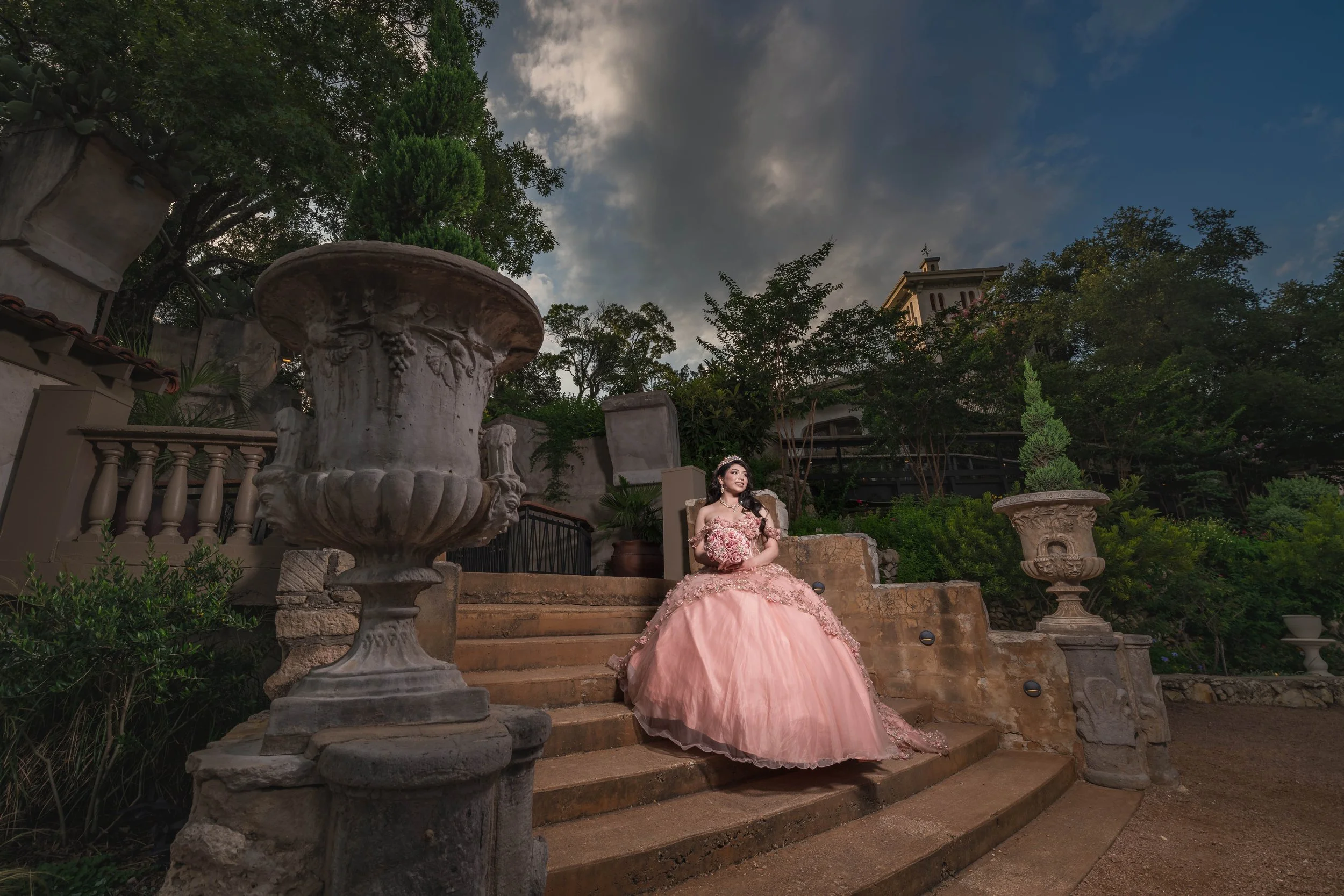 A woman in a pink ball gown sitting on stone stairs outside in a garden, holding a bouquet, with trees and a building in the background during the evening sky.