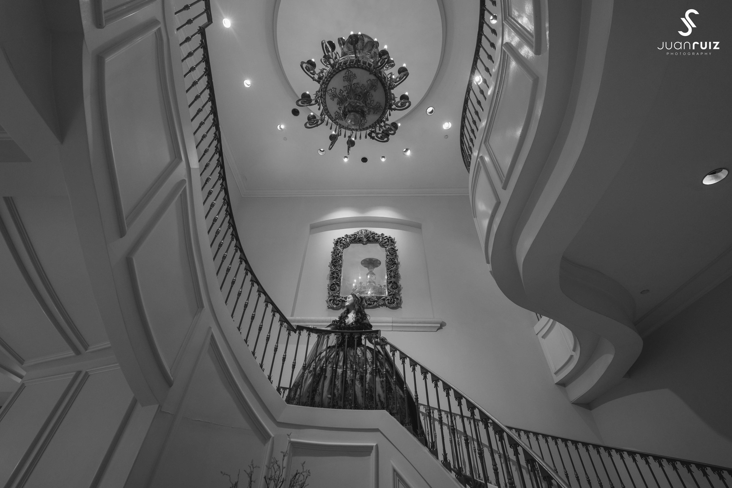 A woman in an elegant gown standing at the top of a grand curved staircase in a luxurious interior with chandeliers and ornate mirror.