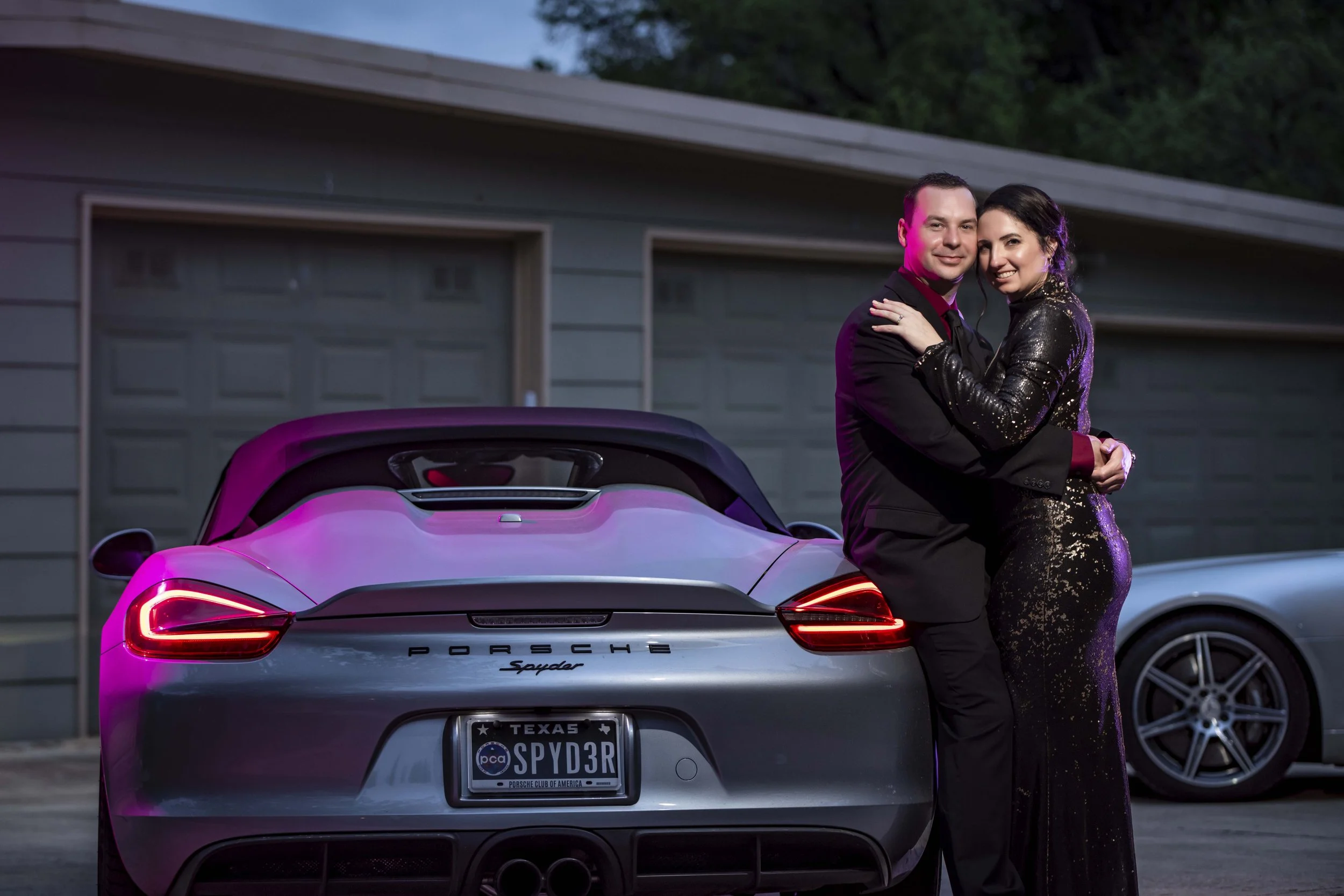 A man and woman dressed in formal attire standing close together next to a silver Porsche Spyder with a Texas license plate that reads 'SPYDR3'. They are outside in front of a garage at dusk, with the woman wearing a black, glittery gown and the man 